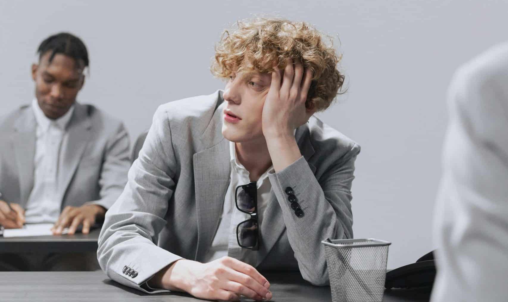 A young man with curly hair, wearing a gray suit, sits at a desk with his head resting on his hand, appearing bored. Meanwhile, another person in the background, sporting a gray suit as well, is writing in a notebook. Perhaps he’s pondering conditions for therapy sessions in Surrey.