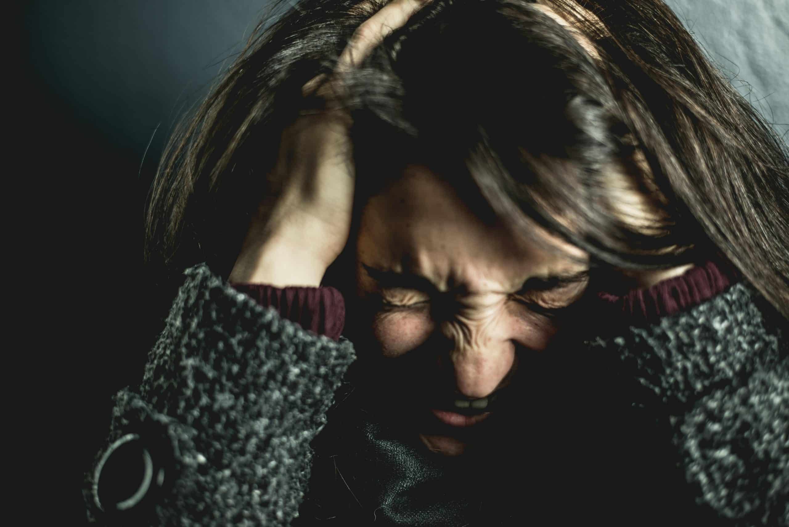 A person with long dark hair clutches their head in distress, eyes closed tightly, wrapped in a dark textured coat. The struggle is all too familiar for many seeking conditions therapy in Surrey, where finding solace and support can be the first step towards healing.