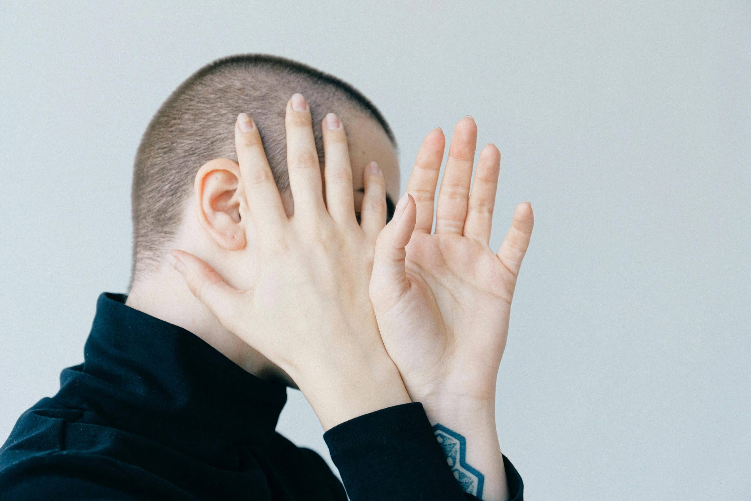 A person with a shaved head covers their face with both hands, perhaps deep in thought about their therapy session. Wearing a black top, they reveal a geometric tattoo on their wrist against the serene backdrop of a plain, light-colored wall in Surrey.